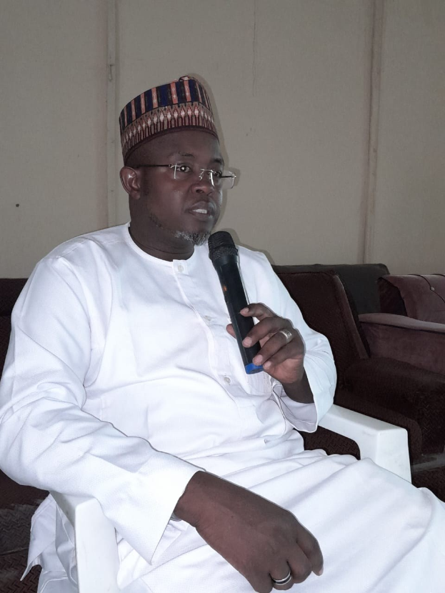 Ein Mann in einem weißen Kleid und einer Mütze sitzt in einem Stuhl und spricht in ein Mikrofon in einem Raum mit zwei Sofas und einer Wand, auf der der Satz "Ich bin kein Lehrer, ich bin ein Lehrer." steht.