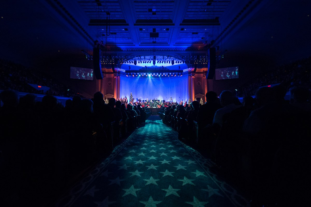 Großer Saal mit sitzendem Publikum, Bühne mit Musikern und Überkopfbeleuchtung und Fernseher, was auf einen Konzertsaal während der Nationalen Convention 2019 hinweist.