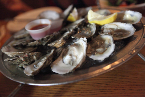 Ein Teller mit Garnelen in einer Reihe, daneben ein Becher mit Sauce und zwei Zitronenscheiben, sowie im Hintergrund sichtbare Teller auf einem Tisch.