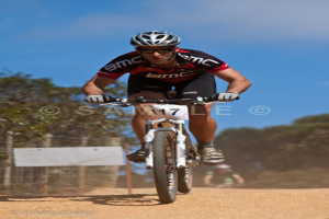 Ein Mann fährt auf einem Fahrrad und trägt ein schwarzes T-Shirt, Shorts und einen Helm, mit Bäumen, Zaun und einem blauen Himmel im Hintergrund und einem Wasserzeichen in der Mitte des Bildes.