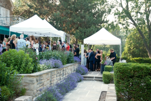 Eine Gruppe von Menschen, die unter weißen Zeltplanen stehen und diskutieren, mit blauen Blumen im Vordergrund und Bäumen im Hintergrund.