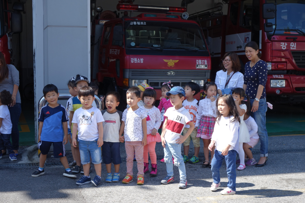 Eine Gruppe von Kindern steht vor einem Feuerwehrauto, einige tragen Mützen, mit mehreren anderen Feuerwehrautos im Hintergrund auf einem Feuerwehrauto.