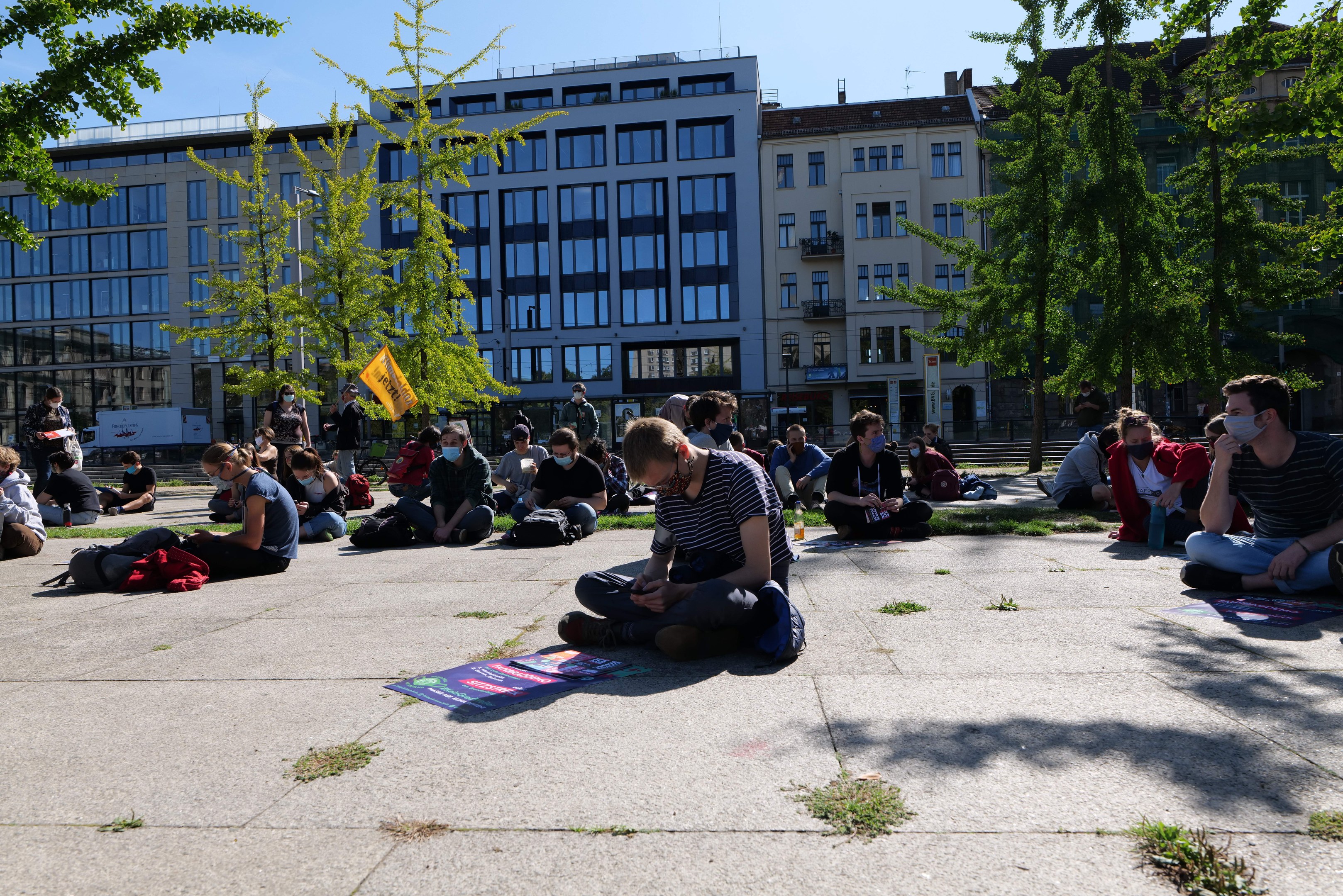 Eine Gruppe von Menschen sitzt vor einem Gebäude auf dem Boden während einer Demonstration in Berlin, umgeben von Bäumen und einem klaren blauen Himmel, wobei einige Masken und Taschen herumliegen.