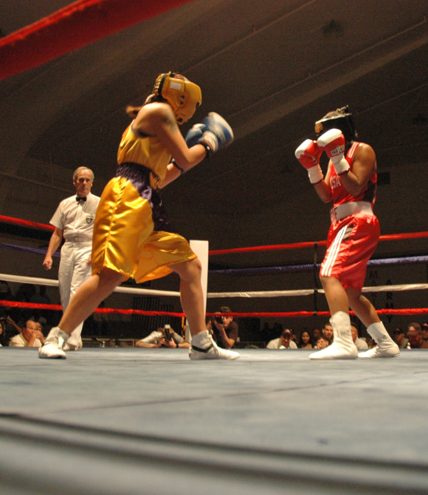 Zwei Boxer im Ring mit einem Schiedsrichter und Zuschauern, einige mit Kameras, unter einer sichtbaren Decke.