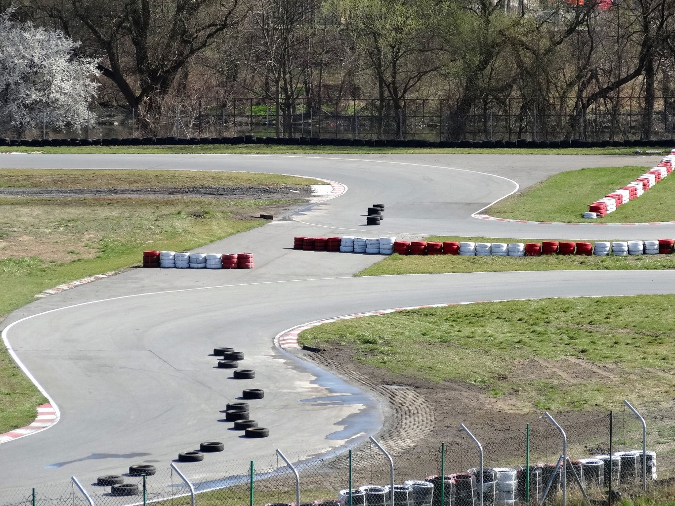 Rennstrecke mit zahlreichen Reifen entlang ihrer Kanten, von Gras und Maschendrahtzaun umgeben, mit Bäumen im Hintergrund.