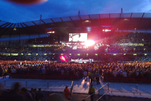 Eine Konzertlocation mit Menschen, Scheinwerfern und einem Projektionsschirm.