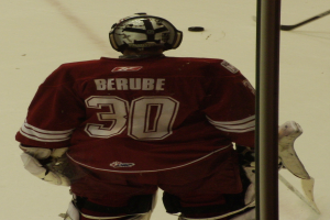 Ein Eishockeyspieler in Helm, Handschuhen und nummeriertem Trikot steht auf dem Eis vor einem Tor mit einem Pfosten rechts daneben.