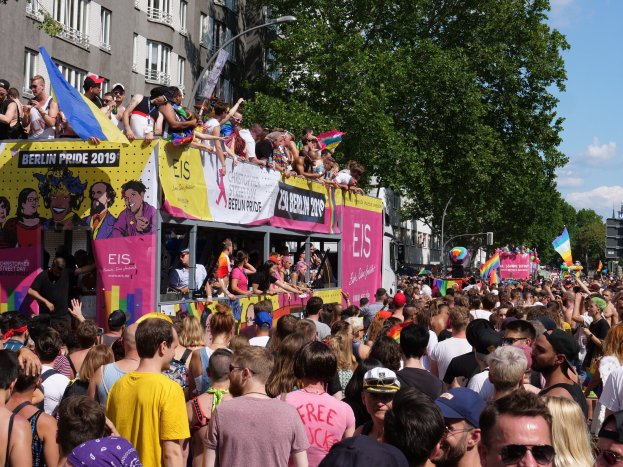 Eine große Menschenmenge marschiert während des Berlin Pride 2019 die Straße entlang, viele tragen Kappen und Goggles, einige halten Fahnen, mit Bannern, Laternenmasten, Bäumen und Gebäuden im Hintergrund unter einem bewölkten Himmel.