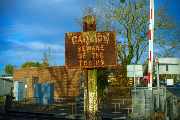 Vorsichtsschild an einem Bahnübergang-Zaun neben Bäumen, Pfählen, einem Gebäude mit Fenstern, Containern, Fahrzeugen auf einer Straße und einem bewölkten Himmel.