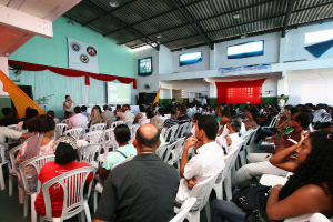 Eine Gruppe von Menschen, die auf Stühlen sitzen und einer Bühne zugewandt sind, auf der ein Polizeibeamter in ein Mikrofon spricht, wobei auf der rechten Seite des Bildes eine Tür zu sehen ist.