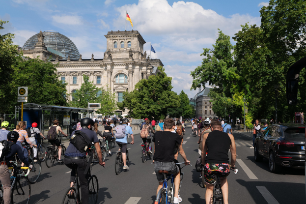 Gruppe von Menschen, die auf Fahrrädern eine von Bäumen gesäumte Straße in Berlin, Deutschland, entlangfahren, mit Gebäuden und einer Bushaltestelle auf der rechten Seite und einem wolkenverhangenen Himmel sowie einer Flagge auf einem Gebäude.