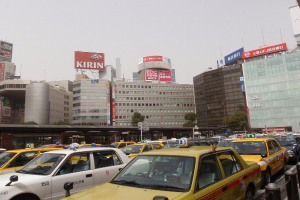 Stadtansicht mit Fahrzeugen auf einer Straße, Gebäuden, Bäumen, Pfählen mit Schildern und Text auf den Schildern, mit einem Zug im Hintergrund und einem Geländer unten rechts.