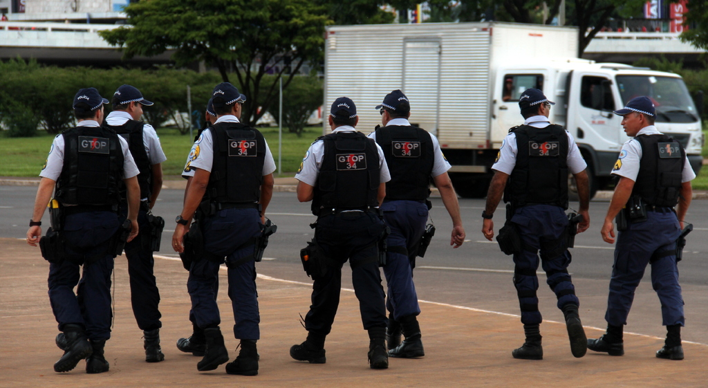 Eine Gruppe von Beamten in Uniform, darunter M√ľtzen, Hemden, Schutzwesten, Uhren, Hosen und Schuhe, die auf einer Stra√Ÿe mit einem geparkten Lkw auf der rechten Seite n√§her an einem Zaun entlanggehen. Im Hintergrund sind Geb√§ude, B√§ume, Pflanzen, Masten, Schilder und eine Br√ľcke in der oberen rechten Ecke zu sehen.