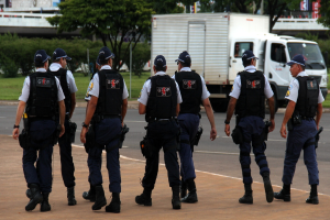 Eine Gruppe von Beamten in Uniform, darunter M√ľtzen, Hemden, Schutzwesten, Uhren, Hosen und Schuhe, die auf einer Stra√Ÿe mit einem geparkten Lkw auf der rechten Seite n√§her an einem Zaun entlanggehen. Im Hintergrund sind Geb√§ude, B√§ume, Pflanzen, Masten, Schilder und eine Br√ľcke in der oberen rechten Ecke zu sehen.