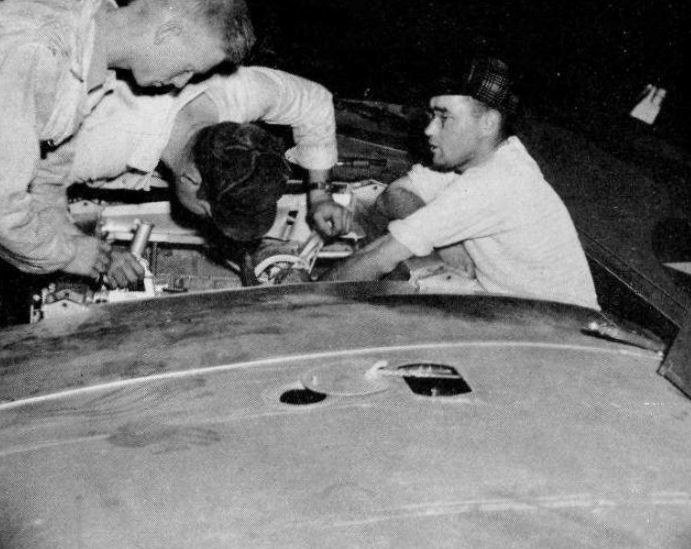 Drei Männer in Mützen reparieren ein schwarz-weißes Auto in einer Werkstatt, umgeben von Werkzeugen und Materialien.