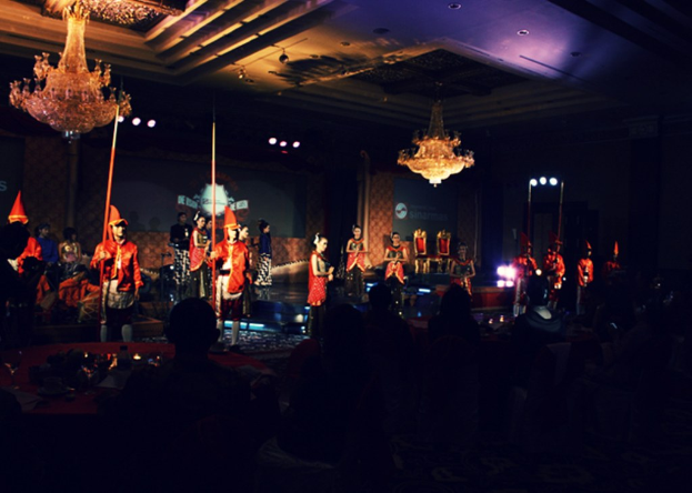 Eine Theateraufführung mit einem Publikum darunter, die zwei an der Decke montierte Kronleuchter zeigt.