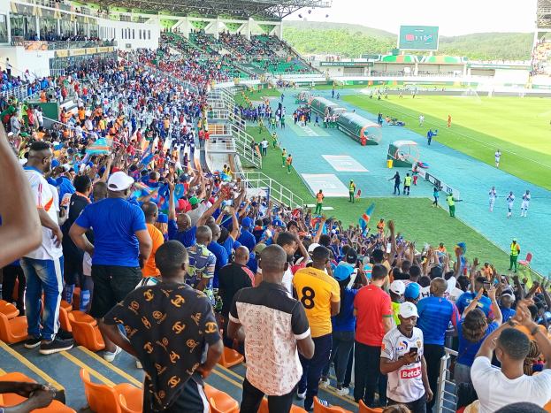 Eine große Menge Menschen versammelt sich vor einem eingezäunten Fußballfeld mit Hügeln und einem klaren Himmel im Hintergrund und erwartet den Beginn eines Spiels während der Afrikameisterschaft 2019.