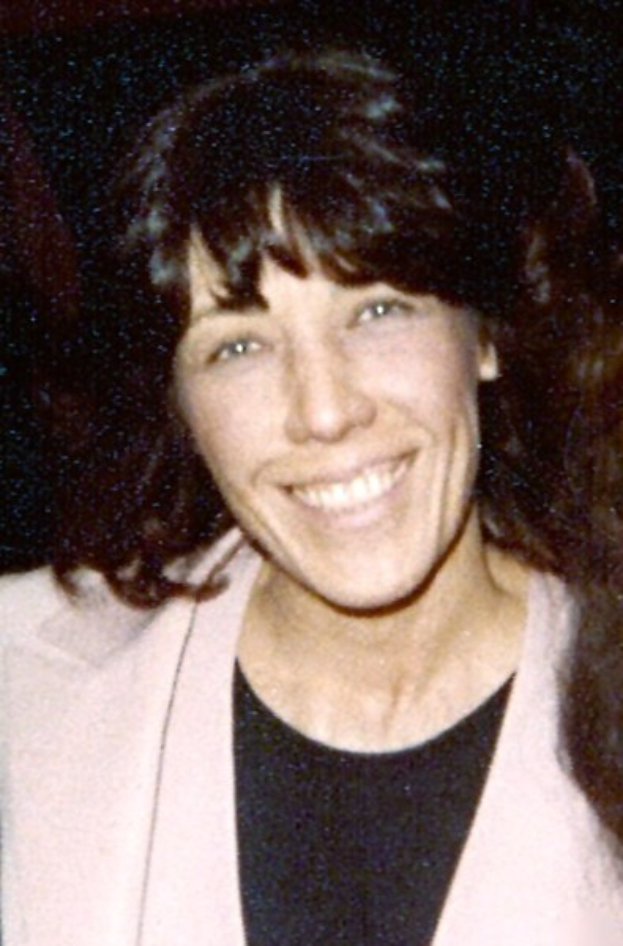 Eine lachende Frau mit langen Haaren, die zu einem Dutt gebunden sind, trägt ein helles Kleid und einen gemusterten Schal vor einem dunklen Hintergrund.