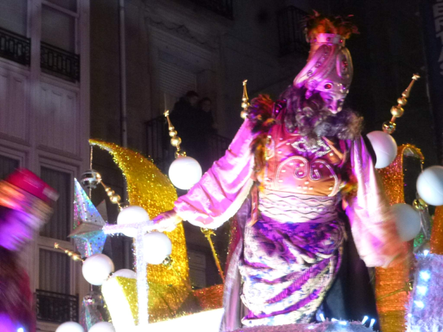 Ein Mann in einem Kostüm steht auf einem dekorierten, beleuchteten Festwagen in einem Stadtumzug, mit einem Gebäude mit Fenstern und Geländern im Hintergrund.