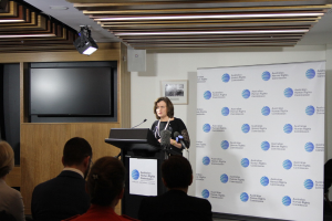 Eine Frau steht auf einer Bühne und spricht in ein Mikrofon, vor ihr sitzen Zuschauer, an der Wand hängt ein Banner, an der Holzwand steht ein Fernseher und an der Decke leuchten Lampen.