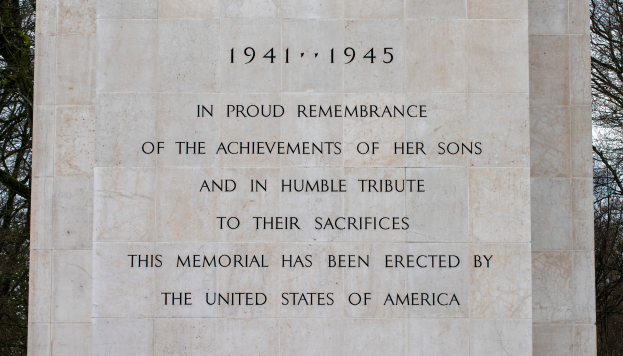 Ein Denkmal mit einer Gedenktafel, die den Text 'In stolzer Erinnerung an die Taten ihrer Söhne und in bescheidener Ehrung ihres Opfers' trägt, vor dem Hintergrund von Bäumen und einem klaren blauen Himmel.