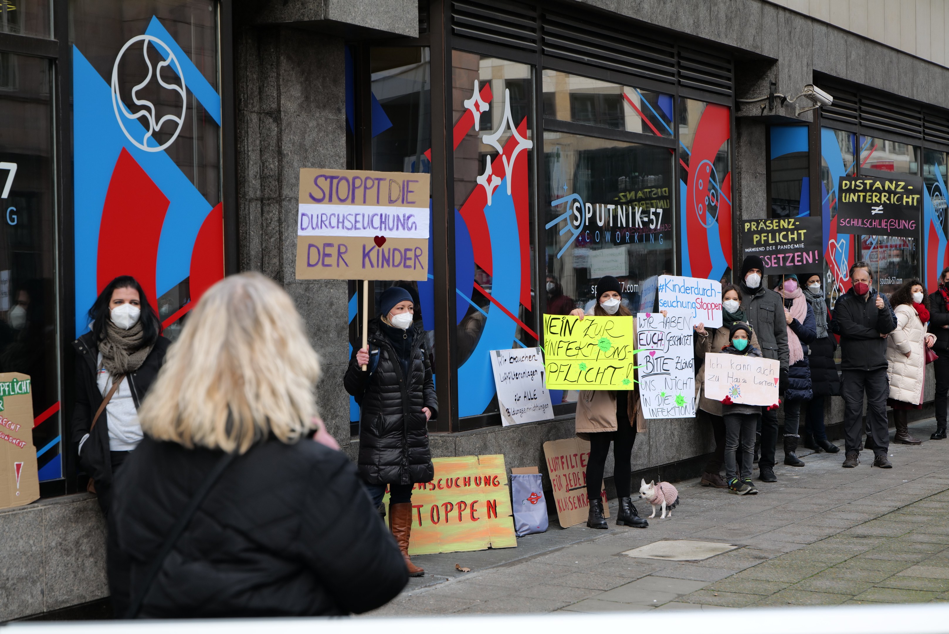 Gruppe von Menschen mit Masken, die Schilder und Plakate vor einem Gebäude mit Glaswänden und -türen halten, mit Kameras und einem Hund anwesend, protestieren gegen eine deutsche Regierungentscheidung, Masken in Schulen zu verbieten.