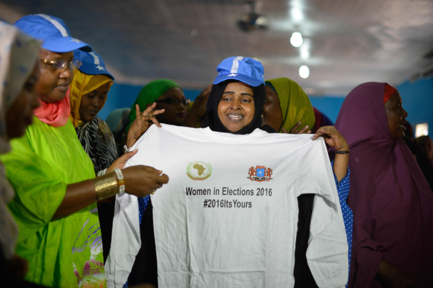 Eine Gruppe von Frauen, die ein weißes T-Shirt mit der Aufschrift "Frauen in Wahlen 2016" hochhalten, einige tragen blaue Mützen, unter Deckenbeleuchtung.