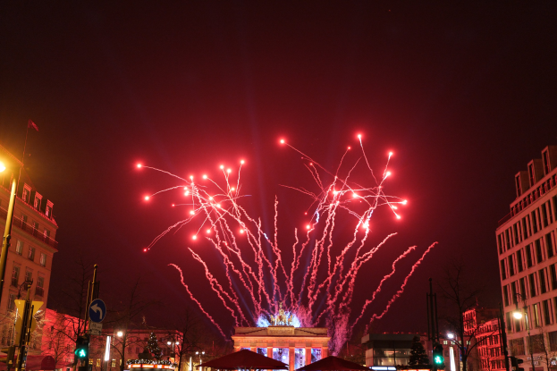 Eine Straßen Szenerie am Silvesterabend in Berlin, mit Gebäuden, Bäumen, Laternen, Ampeln, Schildern, Zelten, Menschen und einem festlichen Feuerwerk am Himmel.