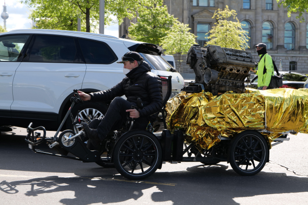 Ein Mann im Rollstuhl mit einem großen Motor am Rücken, umgeben von Fahrzeugen auf einer Straße mit Bäumen, Gebäuden und einem klaren blauen Himmel im Hintergrund.