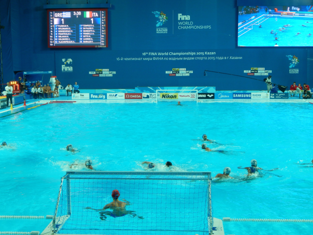 Gruppe von Athleten beim Wasserballspielen in einem Pool während der FINA-Weltmeisterschaften 2016 in Kazan, mit Zuschauern auf der linken Seite und einem Anzeigebrett mit Event-Branding im Hintergrund.