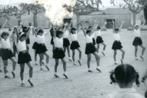Schwarzes und weißes Bild von jungen Mädchen in Schuluniformen, die auf einer Straße tanzen, die von Gebäuden und Bäumen gesäumt ist, mit einem Paar Schuhe am Boden.