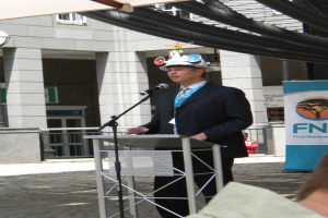 Ein Mann in Anzug und Hut spricht in ein Mikrofon an einem Podium, mit einer Person rechts unten, einem Textband rechts, einem Gebäude und einem Geländer im Hintergrund.