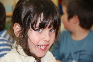 Ein Mädchen in einer Jacke lächelt in die Kamera, während ein Junge in einem blauen T-Shirt wegsieht und eine weitere Person links steht, mit einem unscharfen Hintergrund.