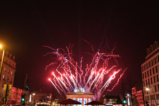 Eine belebte Stadtstraße an Silvester in Berlin, voller Menschen, Fahrzeuge und festlicher D√©korationen, mit Feuerwerk, das den Nachthimmel über den Gebäuden erhellt.