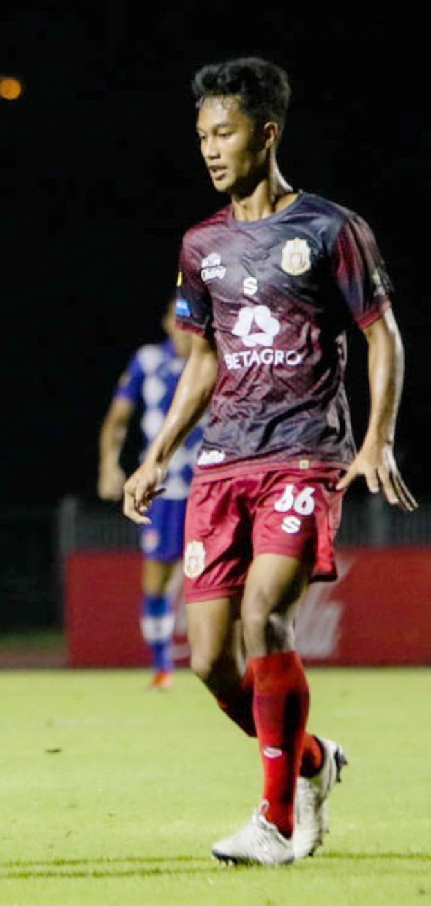 Ein Mann in einem roten Hemd und Shorts spielt Fußball auf einem Feld, mit einer anderen Person im Hintergrund und einer Tafel mit Schrift darauf.