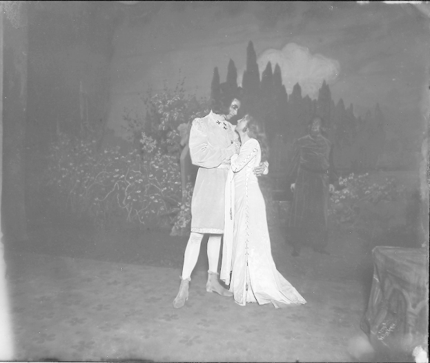 Schwarzes und weißes Foto eines Mannes und einer Frau, die sich auf einer Bühne umarmen und küssen, mit Menschen, Pflanzen und Bäumen im Hintergrund.